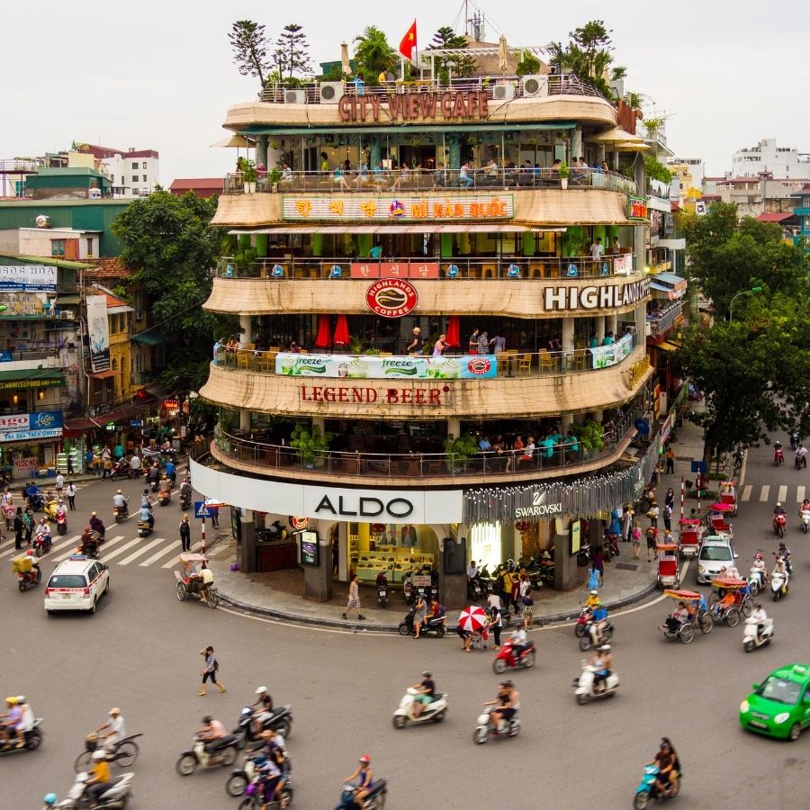 Vivace incrocio a Hanoi, Vietnam, con traffico di scooter e un edificio iconico a più piani con caffè e negozi.