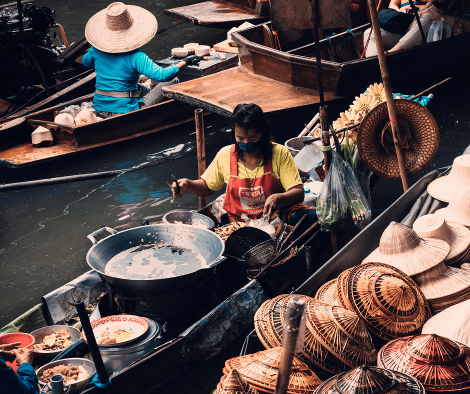 Mercato galleggiante di Phuket: venditrice prepara cibo in barca, cappelli di paglia in primo piano. Esperienza culinaria unica.